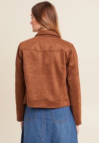 Veste en fausse suède marron avec un col, design ajusté et coutures visibles. Portée sur une jupe en jean, texture lisse.