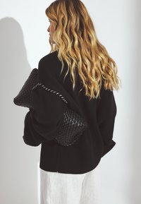 Schwarzer strickpullover mit geripptem Saum, lockere Passform und gewebter schwarzer Clutch-Tasche. Helle Hose vervollständigt das Outfit.