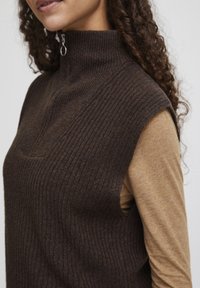 Brun ribbet ærmeløs turtleneck sweater med lynlåsdetalje, lagd over en lys brun langærmet top. Blød tekstur og tætsiddende design.
