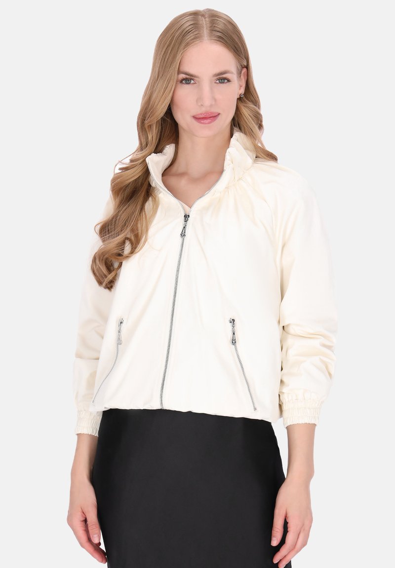 Jachetă crem scurtă, cu guler strâns, două buzunare cu fermoar, manșete elastice și textură netedă a materialului.