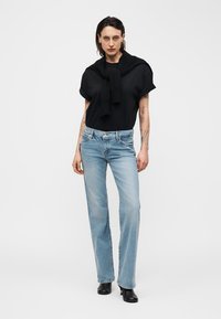 Femme tatouée portant un t-shirt noir et un pull, un jean droit bleu clair et des bottes à talons noires, debout devant un fond uni.