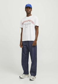 Hvid bomulds t-shirt med grafik og rød tekst samt afslappede mørkeblå denim bukser. Sølv- og grå sneakers med hvide såler. Blå kasket.