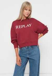 Sweatshirt bordeaux avec le logo "REPLAY" en crème, coupe décontractée, poignets et ourlet en côtes, associé à un jean taille haute bleu clair.