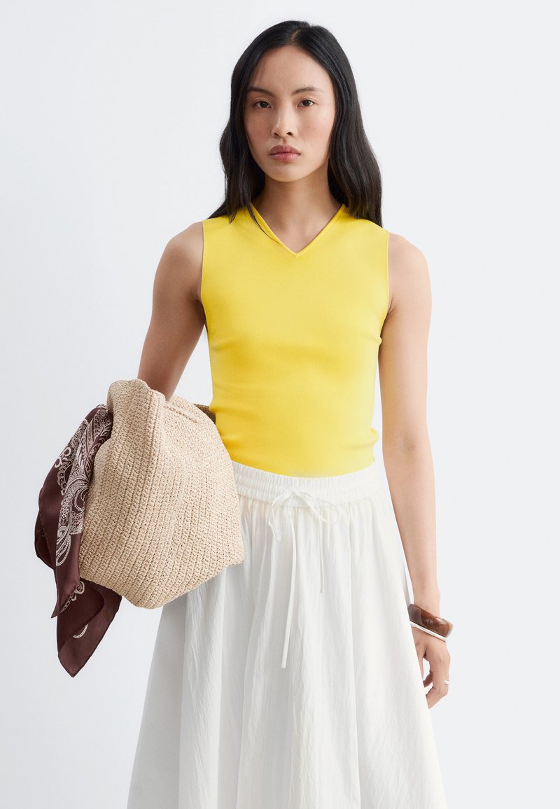 Femme aux longs cheveux noirs portant un haut jaune sans manches et une jupe blanche, tenant un sac beige tissé avec un foulard marron à motifs.