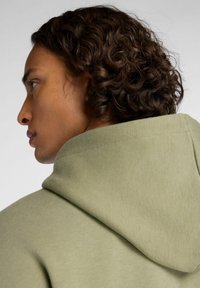 Hoodie in een gedempte groene stof, met een zachte textuur en een relaxte pasvorm. Zichtbare stikdetails langs de rand van de capuchon.
