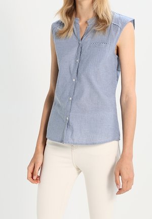 Chemise sans manches bleu clair à boutons, portée avec un pantalon ajusté blanc cassé, présentée sur une personne debout devant un fond uni.