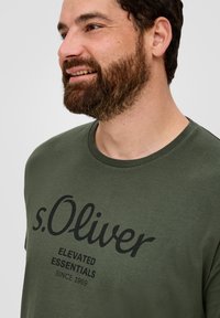 Alyvuogių žalios spalvos medvilnės marškinėliai trumpomis rankovėmis, apvaliu kaklu ir drąsiu juodu tekstu: "s.Oliver ELEVATED ESSENTIALS SINCE 1969" priekyje.