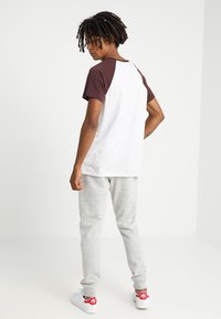 Camiseta raglán blanca y marrón, pantalones de chándal grises, zapatillas blancas con acentos rojos. Modelo visto desde detrás sobre un fondo liso.