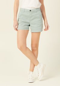 Shorts en coton vert clair avec un devant plat, des passants de ceinture et des poches, associés à des baskets montantes blanches. Texture lisse, design décontracté.