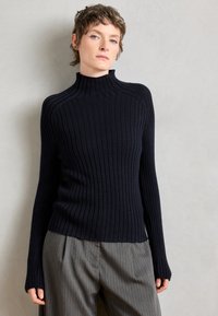 Ribota tumš zilā turtleneck džemperis ar garām piedurknēm un pieguļošu dizainu. Izgatavots no mīksta, teksturēta auduma, apvienots ar gaiši pelēkām svītrainām biksēm.