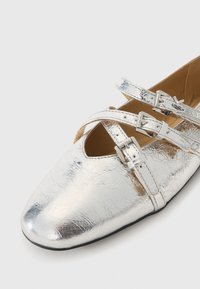 Scarpe piatte metallizzate argento con punta rotonda, dotate di cinturini incrociati fissati da fibbie e una superficie liscia, leggermente strutturata.