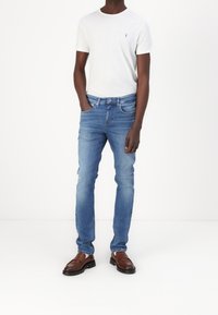 Ljust grå t-shirt med en liten emblem, tillsammans med blå denimjeans och bruna glidskor. Jeansen har en figurnära design och lätt blekning.