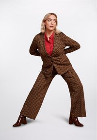 Brun mönstrad kostym med vida byxor och en åtstramad kavaj, lager på lager över en röd skjorta med knappar, tillsammans med bruna klackstövlar.