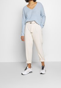 Jasnoniebieski sweter z dekoltem w serek, miękki materiał; kremowe spodnie zwężane z paskiem do wiązania; białe sneakersy z czarnymi i złotymi akcentami.