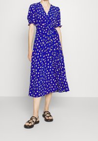 Blue wrap dress with short puff sleeves, featuring a scattered floral pattern in white and yellow. Worn with black strappy sandals.