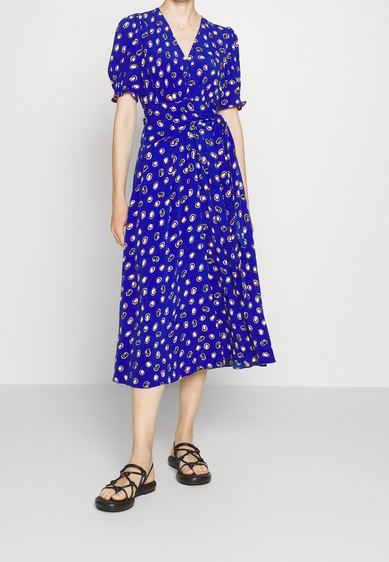 Blue wrap dress with short puff sleeves, featuring a scattered floral pattern in white and yellow. Worn with black strappy sandals.