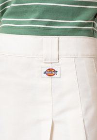 Bílé kalhoty Dickies mají hladkou tkaninu, rovný střih, záhyby vpředu a logo na zadním pásku.