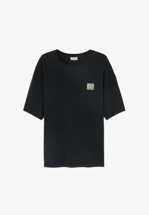 Melna kokvilnas T-kreklā ar īsām piedurknēm, apaļo kakla izgriezumu un mazu baltu kvadrātveida logo, kas priekšā rakstīts "AMERICAN VINTAGE".