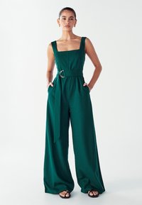 Mujer de pie que lleva un mono sin mangas de color verde oscuro con cinturón y sandalias negras, con las manos en los bolsillos, contra un fondo blanco.