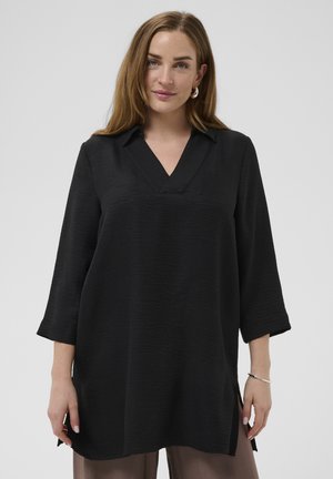 Femeie cu păr lung și maro, purtând o tunică neagră lejeră cu decolteu în V și mâneci trei sferturi, și pantaloni maro, stând pe un fundal simplu.