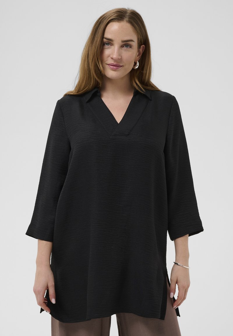 Femme aux longs cheveux bruns portant une tunique noire à col en V et manches trois-quarts, ainsi qu'un pantalon marron, debout devant un fond uni.