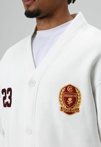 Witte button-up cardigan met een geborduurd bordeauxrood embleem en het nummer "23" aan de voorkant, met een zachte textuur en een sjaalkraag.