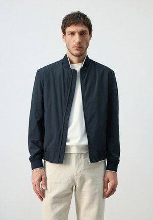 Hombre con chaqueta azul marino con cremallera sobre un suéter blanco y pantalones beige, de pie contra un fondo liso de color gris claro.