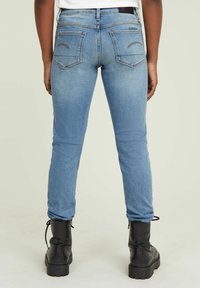 Person klädd i ljusblå, tighta jeans och svarta snörstövlar, stående med ryggen vänd mot kameran mot en neutral bakgrund.