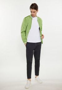 Bombardiere verde chiaro, t-shirt bianca, pantaloni neri a sigaretta e sneakers bianche; la giacca presenta una chiusura con zip e colletto a coste.