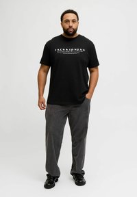 Czarny bawełniany t-shirt z białym logo, zestawiony z szarymi spodniami sztruksowymi. Model stoi ze wzrokiem w dół, z rękami w kieszeniach, w czarnych butach.
