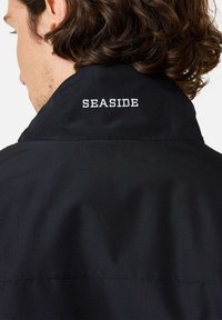Giacca nera con colletto alto e testo ricamato "SEASIDE" sul retro. Il materiale presenta una delicata trama testurizzata.