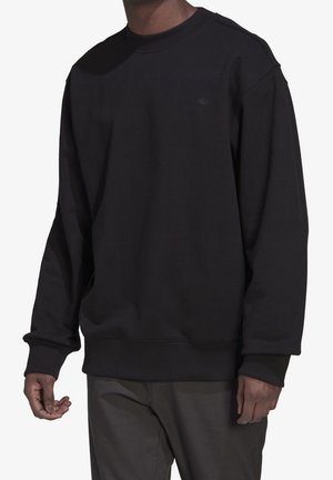 Mann trägt schlichtes schwarzes Sweatshirt mit Rundhalsausschnitt und dezentem Logo auf der Brust sowie dunkle, strukturierte Hose, steht vor weißem Hintergrund.