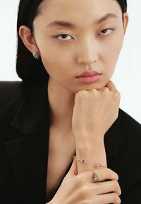 Pulsera de oro con dos piedras rosas, pendientes de plata a juego con acentos azules. El modelo tiene una expresión neutral y lleva un blazer negro.