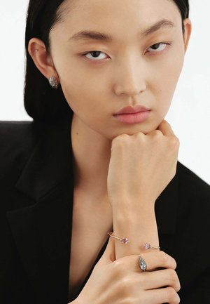 Pulsera de oro con dos piedras rosas, pendientes de plata a juego con acentos azules. El modelo tiene una expresión neutral y lleva un blazer negro.