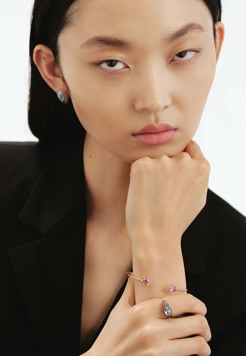 Pulsera de oro con dos piedras rosas, pendientes de plata a juego con acentos azules. El modelo tiene una expresión neutral y lleva un blazer negro.