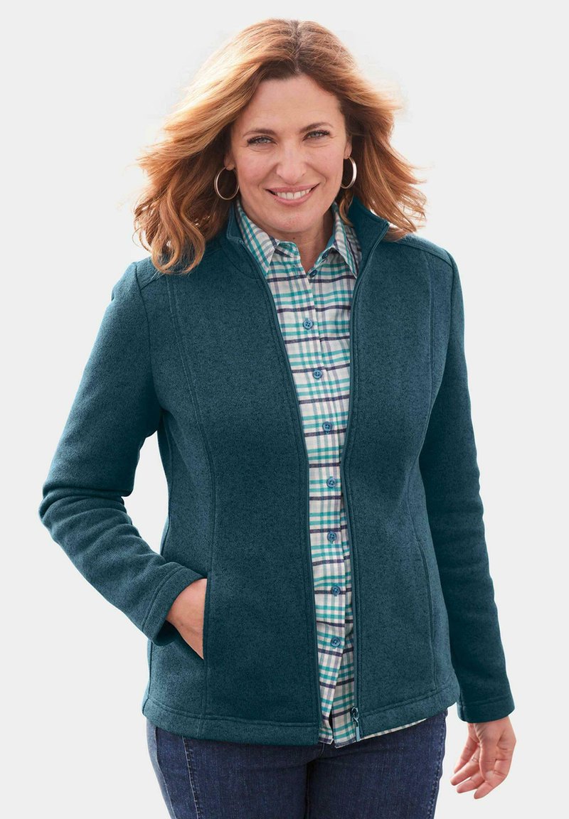 Türkiser Fleece-Jacke mit hohem Kragen, ausgestattet mit einem Reißverschluss und seitlichen Taschen. Getragen über einem blau-grünen Karoshirt.