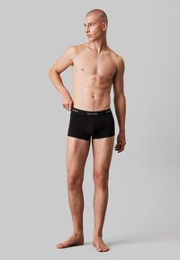 Black cotton boxer shorts with elastic waistband featuring "Calvin Klein" branding, fitted design, and smooth texture. Model standing barefoot.