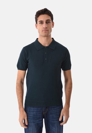 Giovane uomo in piedi, indossa una polo a maniche corte blu scuro con tre bottoni e jeans blu, fronteggiando davanti a uno sfondo semplice.