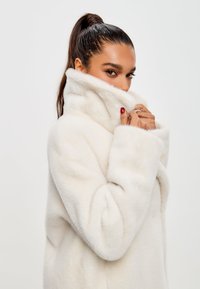 Weißer Kurzhaarmantel aus Kunstfell mit hohem Kragen und weiten Ärmeln, der eine weiche Textur aufweist. Das Model hält den Kragen nah ans Gesicht.