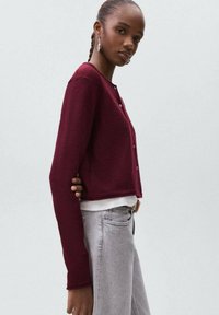 Burgundy kort cardigan med rund halsringning, framknappar och långa ärmar; bärs över en vit topp och matchas med ljusgrå jeans.