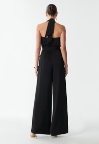 Woman with hair in bun wears black halter-neck jumpsuit with wide legs and gold high-heeled sandals, standing against a white background.