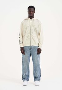 Sudadera con cremallera de color beige claro con diseño gráfico de calavera, cordón ajustable y acentos de rayas negras en las mangas; combinada con jeans azules holgados.