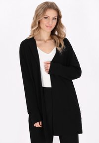 Cardigan nero realizzato in tessuto morbido, con frontale aperto, maniche lunghe e vestibilità morbida. Indossato sopra una maglietta bianca per creare contrasto.