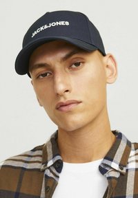 Navy blue cap with white "JACK&JONES" logo, cotton material, curved brim, worn with a plaid shirt, featuring brown, white, and navy colors.