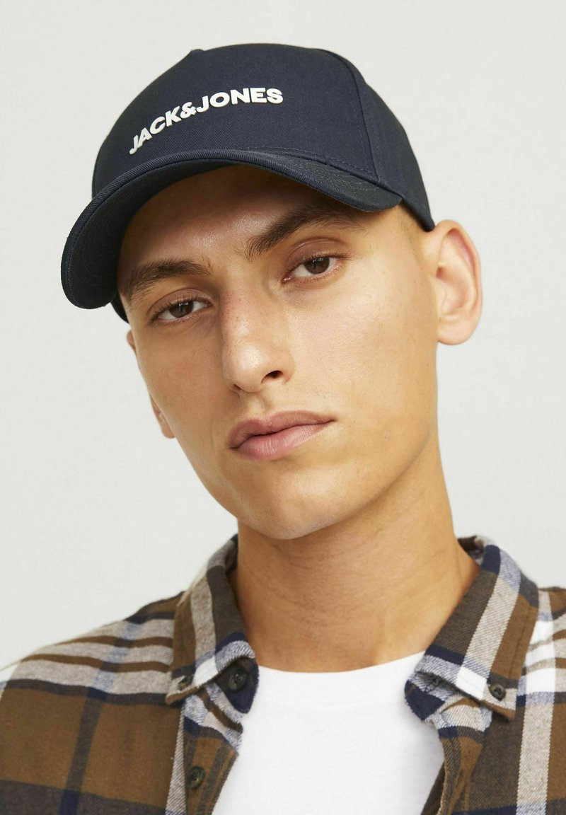 Navy blue cap with white "JACK&JONES" logo, cotton material, curved brim, worn with a plaid shirt, featuring brown, white, and navy colors.