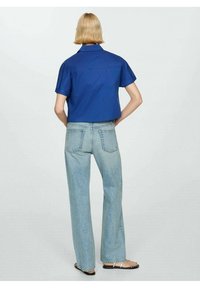 Camicetta blu corta con maniche corte, dotata di colletto, abbinata a jeans a zampa d'elefante chiari e sandali con cinturini.