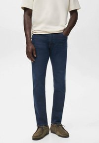 Hombre vistiendo jeans slim-fit de color azul oscuro, una camisa beige de manga corta y zapatos casuales marrones con cordones, con una mano en el bolsillo.
