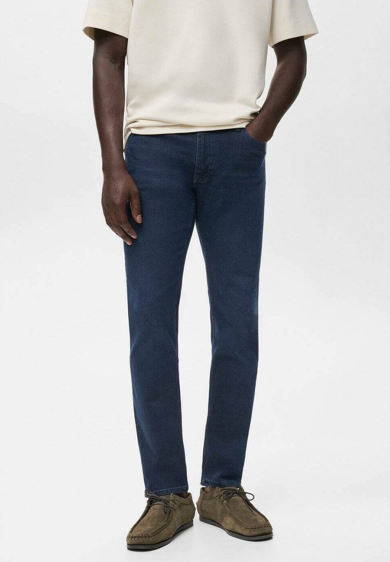 Hombre vistiendo jeans slim-fit de color azul oscuro, una camisa beige de manga corta y zapatos casuales marrones con cordones, con una mano en el bolsillo.