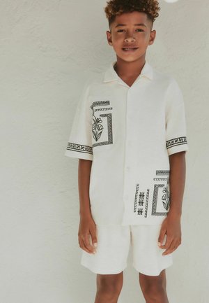Boy with curly hair wearing a white short-sleeve shirt and shorts set with black geometric and floral patterns, standing against a light wall.