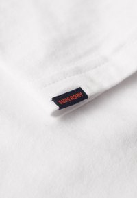 Biała bawełniana koszulka z zagiętym kołnierzykiem i czarną metką z napisem "SUPERDRY" w pomarańczowym kolorze po boku. Miękka tekstura materiału.